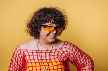 Güneş gözlüklü ve eşsiz giyinen mutlu insan sarı arka planda neşe ve bireysellik sergiliyor.