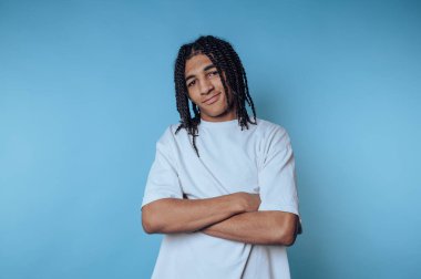 Saçları örgülü şık genç adam, modern ve havalı bir hava sergileyen parlak mavi bir arka planda kendine güvenerek poz veriyor.