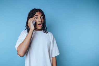 Rastalı genç adam, mavi arka planda şaşkınlık ve şaşkınlık gösteren akıllı bir telefonla konuşuyor.