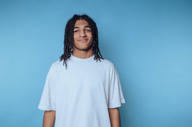 Gülümseyen genç adam mavi bir duvara sıcak bir şekilde karşı duruyor, pozitifliği ve enerjiyi somutlaştırıyor.