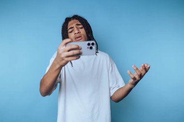 Genç adam telefonuna bakarken hayal kırıklığını ifade ediyor. Mavi arka planda hayal kırıklığı ve duygu dolu bir anı yakalıyor.