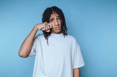 Genç insan derin düşüncelere dalmış bir ifade sergiliyor, belirsizlik ve endişeyi, mavi arka planda düşünceli bir duruşla tasvir ediyor.