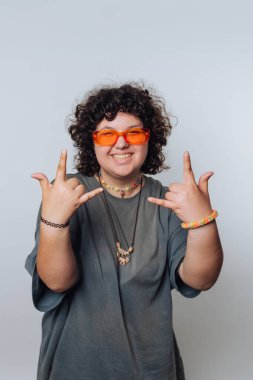 Neşeli el işaretleri ve şık güneş gözlükleriyle gülümseyen genç bir şahıs, iç mekan ortamında hafif bir arka planda poz veriyor.