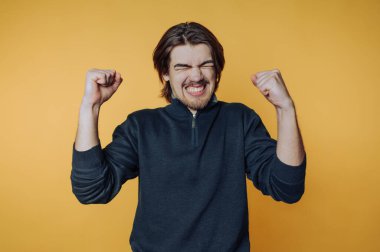 Genç adam zaferi büyük bir gülümsemeyle kutluyor ve parlak sarı bir arka plana karşı yumruklarını kaldırıyor.