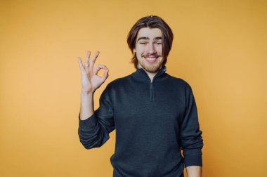 Genç adam, parlak sarı bir arka plana karşı neşeli bir ifadeyle iyi bir jest yapıyor.