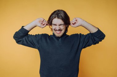 Kapalı alandaki eğlenceli anlarda parlak sarı arkaplan önünde şakacı kulakları tıkayan genç bir adam.