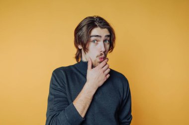 Genç adam çenesine dokunurken parlak sarı bir arka plana karşı düşünceli bir ifade takınıyor ve meraklı görünüyor.