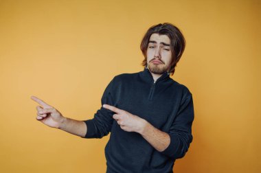 Şık saçlı genç adam canlı sarı bir arka planda poz veriyor, iki eliyle şakacı hareketler yapıyor.