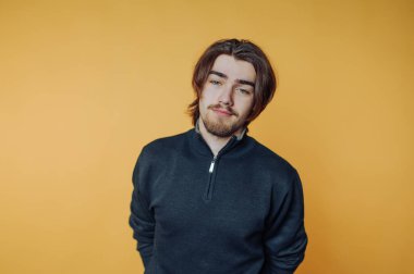 Genç adam, sıradan bir iç mekan ortamında parlak sarı bir arka plan önünde güven içinde poz veriyor.