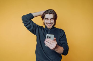 Genç adam, parlak sarı arka planda akıllı telefonuna bakarken gülümsüyor.