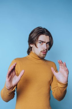 Genç adam, zarif bir hardal balıkçı yaka kazak giyerken mavi bir arka planda elleri olan hareketler yapıyor.