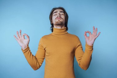 Genç adam, açık mavi bir arka planda, rahat bir balıkçı yaka kazak giyerken sakince meditasyon yapıyor.