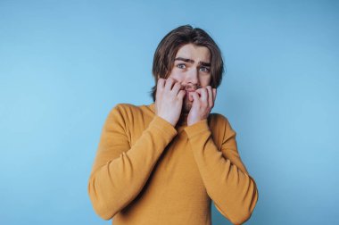 Genç adam, bir stüdyo ortamında sıradan bir süveterle mavi bir arka plana karşı endişe ve korku ifade ediyor.