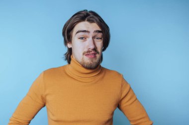 Turuncu balıkçı yaka kazak giyen genç adam açık mavi bir arka plana karşı gevşek bir ifadeyle poz veriyor.