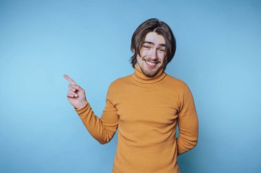 Balıkçı yaka kazaklı genç adam, içten bir şekilde gülümserken mavi bir arka plana şakacı bir şekilde işaret ediyor.