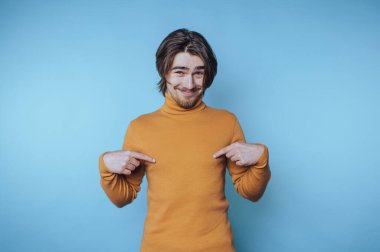 Boğazlı kazaklı gülümseyen genç adam mavi bir arka plana doğru hareketler yapıyor.