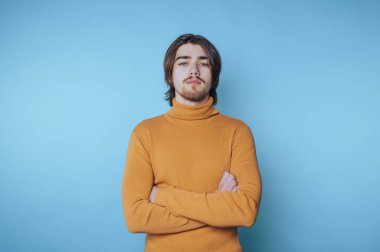 Sarı balıkçı yaka kazaklı, kendine güvenen genç adam, kolları kapalı fotoşopta çapraz duran mavi arka planda duruyor.