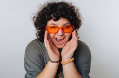 Kıvırcık saçlı olan kişi turuncu güneş gözlüğü takıyor ve beyaz bir fonda poz verirken sevinçle gülümsüyor.