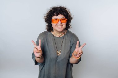 Kıvırcık saçlı genç adam, turuncu güneş gözlüğü ve sıradan bir kıyafetle stüdyoda sade bir arkaplanda poz veriyor.