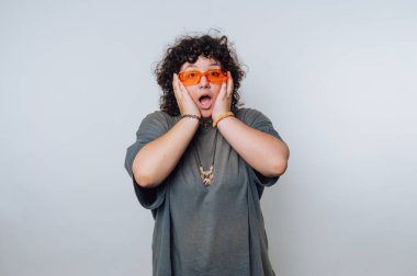 Turuncu güneş gözlüğü takan kıvırcık saçlı heyecanlı biri açık bir arkaplan karşısında şaşırmış görünüyor.