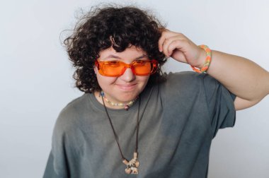Turuncu güneş gözlüğü takan kıvırcık saçlı genç, saçlarına dokunurken arka planda rahatça poz veriyor.