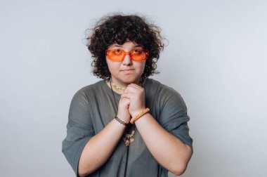 Kıvırcık saçlı, turuncu gözlüklü ve aksesuar takan biri elleri düz bir arka plana yapıştırılmış şekilde poz verir.