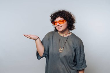 Kıvırcık saçlı ve turuncu güneş gözlüklü genç bir yetişkin, tek eliyle hareket ederken tarafsız bir arkaplana güvenle poz verir.