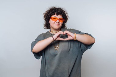 Büyük tişörtlü genç adam elleriyle kalp hareketi yapıyor, stüdyo ortamında turuncu güneş gözlüğü takıyor.