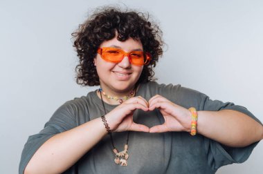 Elleriyle kalp işareti gösteren, güneş gözlüğü takan ve renkli aksesuarlar takan genç bir adam.