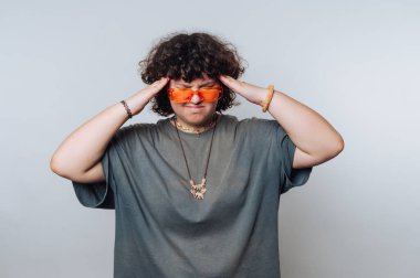 Turuncu güneş gözlüğü takan kişi, basit bir stüdyo ortamında ciddi görünürken başını tutuyor.