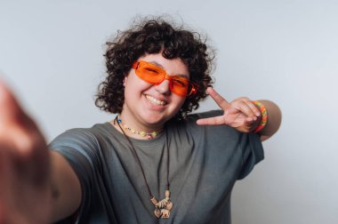 Kıvırcık saçlı, turuncu gözlüklü, gülümseyen ve iç mekan ortamında barış işareti ile poz veren biri.