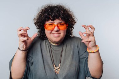 Kıvırcık saçlı ve turuncu gözlüklü kişi, neşeli bir jest, gösteriş tarzı ve kapalı alanda kendine güveni gösterir.
