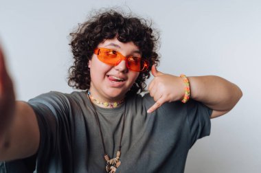 Genç insan renkli aksesuarlar ve kendine özgü güneş gözlükleriyle evin içinde selfie çekerken neşeli bir jest yapıyor.