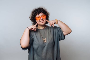 Kıvırcık saçlı, parlak turuncu güneş gözlüklü genç, açık bir arkaplanda şakacı bir poz veriyor.