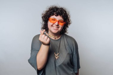 Kıvırcık saçlı genç kalp hareketi yapıyor, sade arka planda turuncu güneş gözlüğü takıyor.