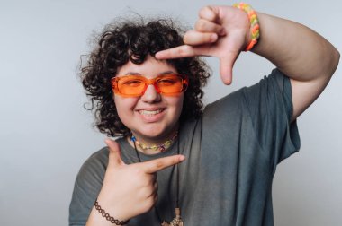 Kıvırcık saçlı genç adam gündüz vakti stüdyoda parlak gözlük takarken neşeli bir jest yapıyor.