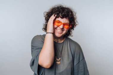 Kıvırcık saçlı, turuncu gözlüklü biri yaratıcı bir ortamda ışık perdesine karşı şakacı bir şekilde poz verir.