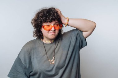 Kıvırcık saçlı ve turuncu gözlüklü biri kafasına dokunurken tuhaf bir yüz ifadesi sergiliyor. Benzersiz bir moda anlayışı ve oyuncu bir hava sergiliyor.