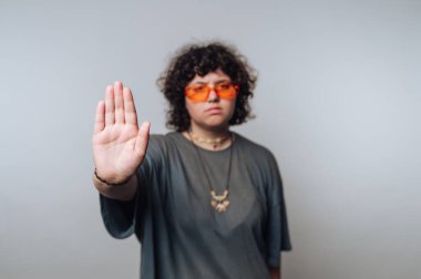Kıvırcık saçlı, turuncu güneş gözlüklü biri, düz bir arkaplan karşısında durarak el kaldırıyor.