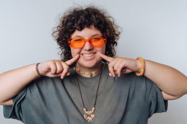 Kıvırcık saçlı, turuncu gözlüklü, gülümseyen ve yanaklarını gösteren, açık renk aksesuarlar takan genç bir adam.