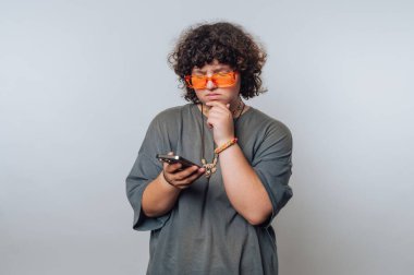 Nötr arkaplana sahip minimalist alanda düşünürken telefonu tutan düşünceli bir genç.