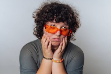 Kıvırcık saçlı, turuncu güneş gözlüğü takıp, iç mekanda günlük kıyafet giyerken dalgın duygular gösteren etkileyici bir birey.