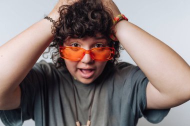 Kıvırcık saçlı, turuncu gözlüklü, elleri kafasının üstünde, açık bir arkaplanda sürpriz yapan biri.