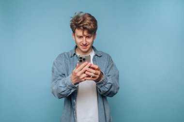 Genç adam akıllı telefonuyla sıradan bir ortamda saf mavi bir arkaplan üzerinde etkileşim kuruyor.