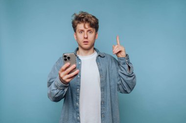 Telefonlu bir adam, sıradan bir ortamda mavi bir arkaplanda dururken şaşkınlığını gösteriyor.