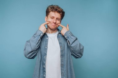 Genç adam neşeli bir şekilde mavi bir arkaplan karşısında komik bir ifade sergiliyor.