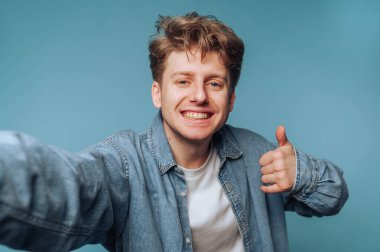 Mutlu genç adam, sıradan bir ortamda mavi bir arkaplan karşısında selfie çekerken başparmağını kaldırıyor.