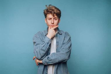 Saçları dağınık genç bir adam. Düşünceli bir şekilde mavi bir arkaplana karşı poz veriyor.