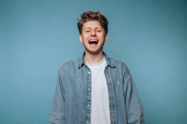 Sıradan giyinmiş gülümseyen genç adam mavi arka planda eğleniyor.