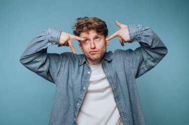 Genç adam mavi arka planda şakacı poz veriyor, elleriyle komik hareketler yapıyor.